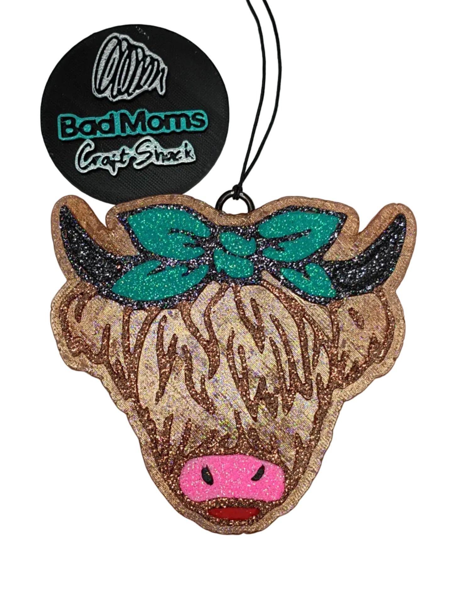 Highland cow shaped car freshener-with raised details. Rose gold glitter hair with black glitter horns, a pink glitter nose, red glitter tongue and a handkercheif style bow in teal glitter outlined in black glitter on a broze brown base.