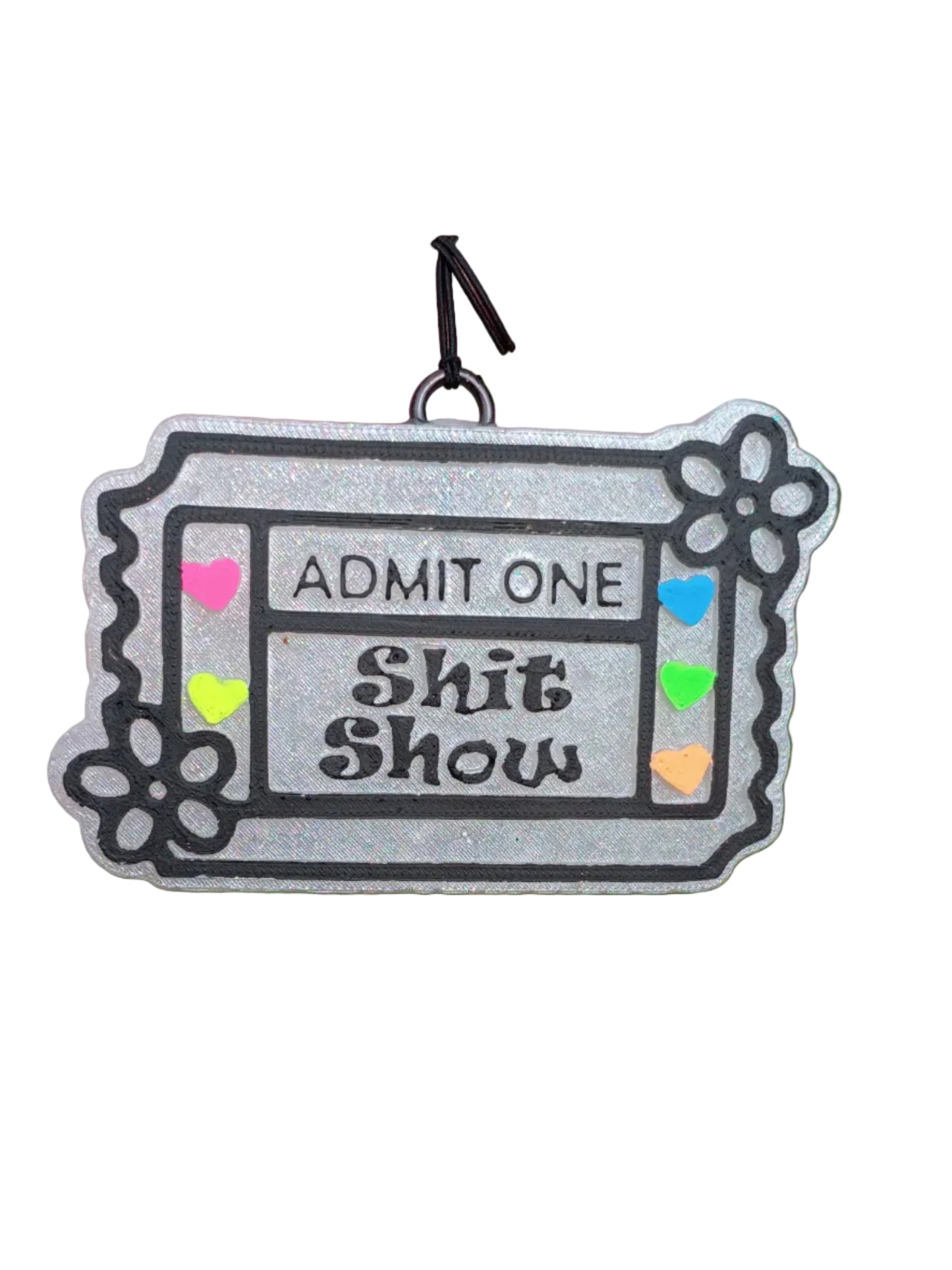 Ticket shaped base in pearl white. Black lettering says ADMIT ONE SHIT SHOW in the middle. Raised classic ticket lines outlined in black paint. Raised daisy flower on two corners outlined in black paint. Neon painted hearts in pink, orange, yellow, green and blue on the 2 sides of the wording.