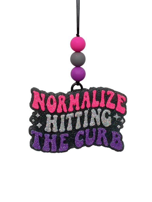 Stacked vertically the words Normalize Hitting The Curb in hot pink, silver and purple glitter lettering on a black base.  Three silicone beads in pink, grey and purple decorate the black elastic hanger. 