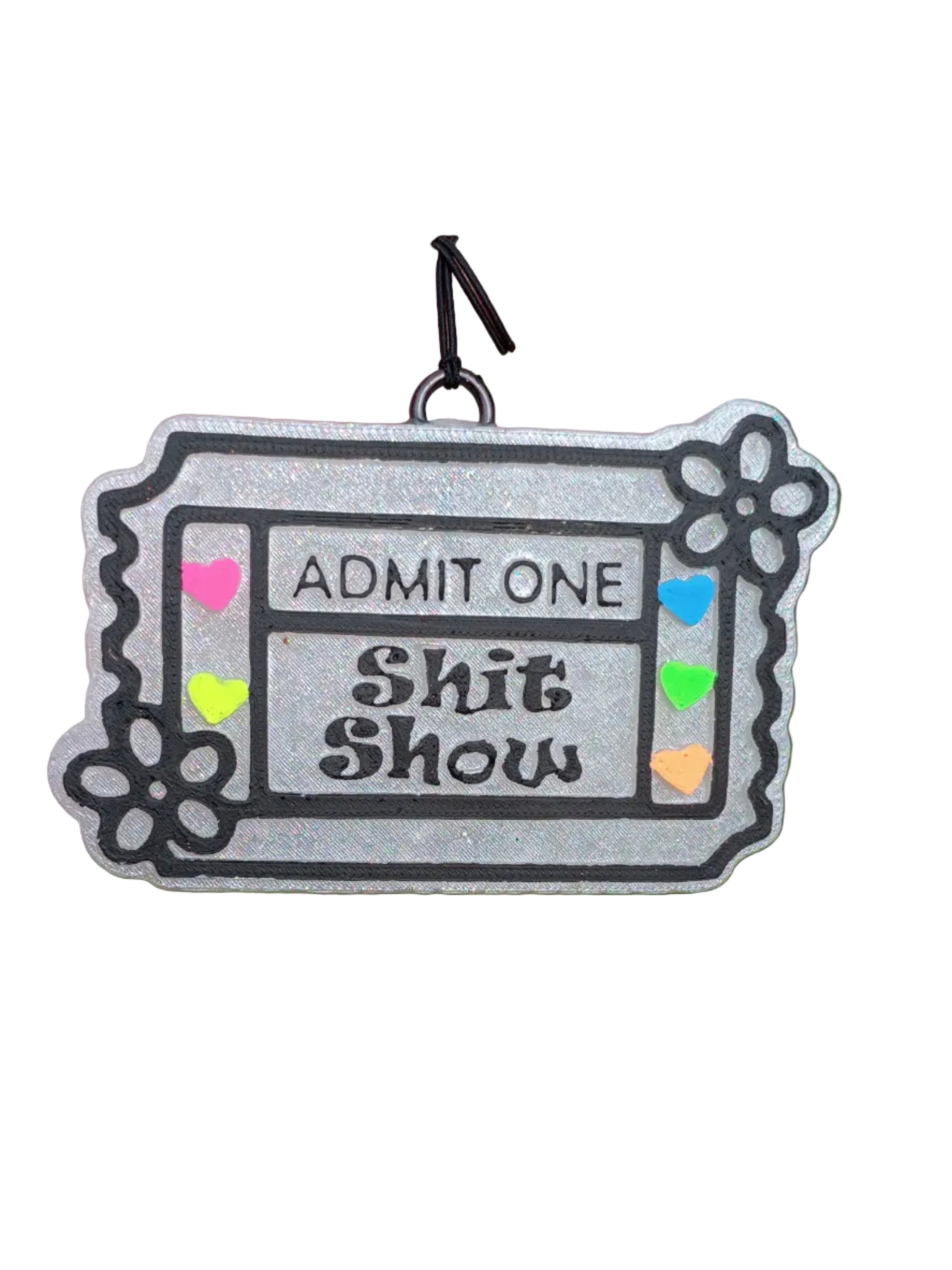 Ticket shaped base in pearl white. Black lettering says ADMIT ONE SHIT SHOW in the middle. Raised classic ticket lines outlined in black paint. Raised daisy flower on two corners outlined in black paint. Neon painted hearts  in pink, orange, yellow, green and blue on the 2 sides of the wording. 