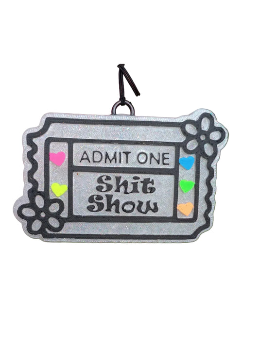 Ticket shaped base in pearl white. Black lettering says ADMIT ONE SHIT SHOW in the middle. Raised classic ticket lines outlined in black paint. Raised daisy flower on two corners outlined in black paint. Neon painted hearts  in pink, orange, yellow, green and blue on the 2 sides of the wording. 