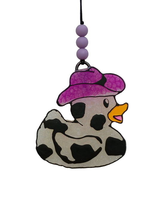 Duck shaped car freshener with black cow print wearing a purple cowboy hat on a white base. Purple beads decorate the elastic hanger. 