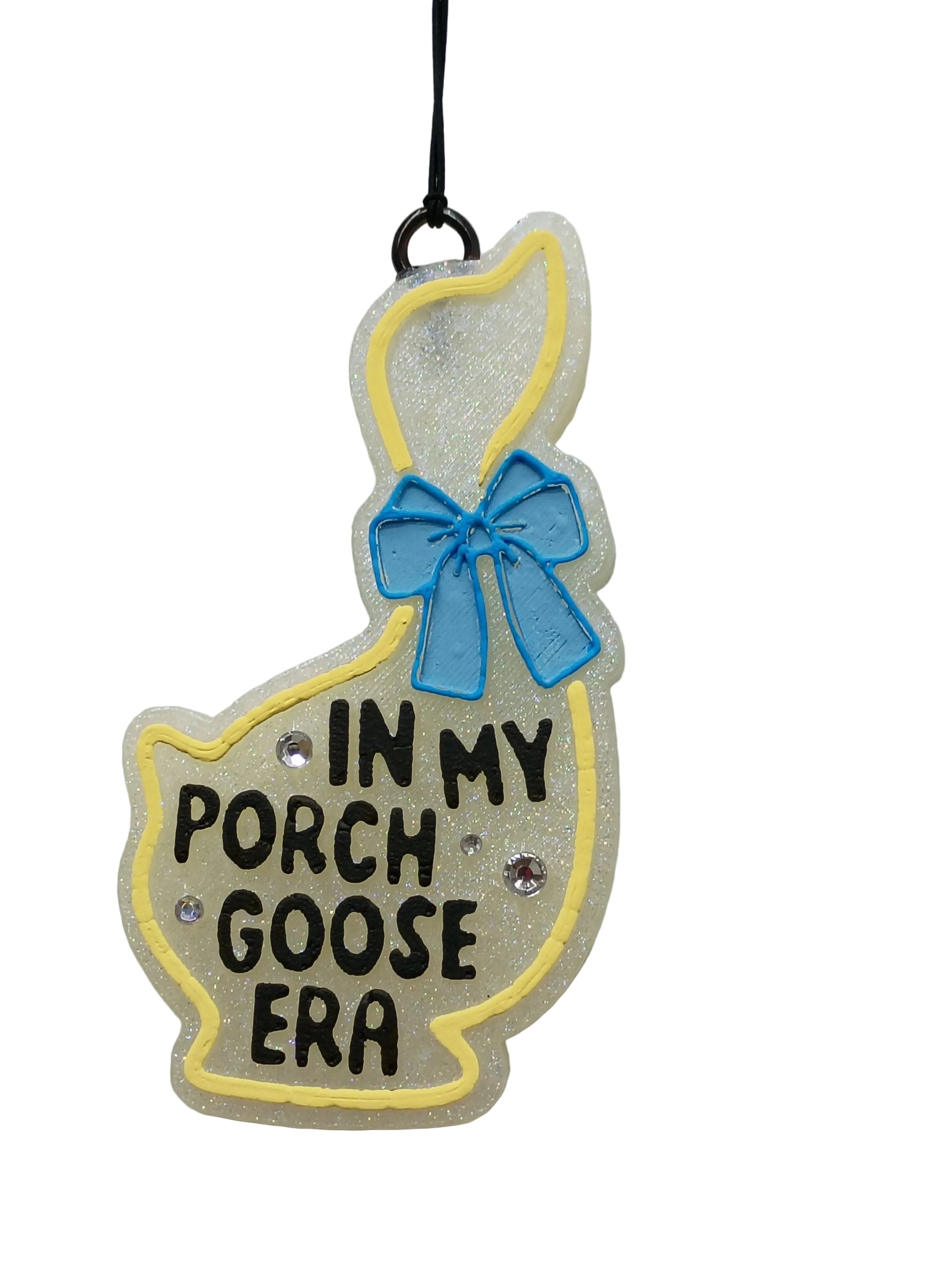 Decorative goose shaped car freshener  with 'In My Porch Goose Era' text  in black , a blue bow and yellow outline around the goose on a white base. 3 rhinestones scattered around the lettering.